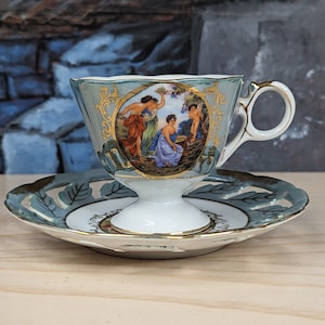 May include: A delicate porcelain teacup and saucer set with a blue and gold floral design. The cup features a scene of three women in a garden setting.