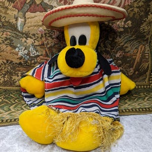 May include: A plush yellow dog wearing a sombrero and a colorful striped poncho. The dog has black ears, a black nose, and a white and red hat. The poncho has red, blue, green, and yellow stripes.