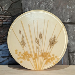 May include: A round, white plate with a fan-shaped design of dried grass and spaghetti noodles. The plate is decorated with small, yellow pasta rings.