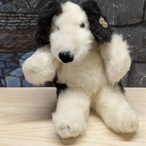 May include: A plush dog toy with a black and white pattern. The toy has black ears and a black nose. The dog is sitting on a wooden surface. A small decorative element is attached to one ear.