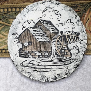 May include: A round, decorative plate featuring a detailed relief of a water mill scene. The plate has a textured, cracked white border and a central image in shades of brown and black. The water wheel and mill building are prominent.