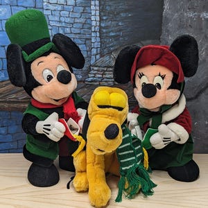 May include: Three plush toys featuring Mickey Mouse wearing a green hat and vest, Minnie Mouse in a red bonnet and green dress, and Pluto wearing a green and white striped scarf. The characters are posed together with soft, textured fabric details.