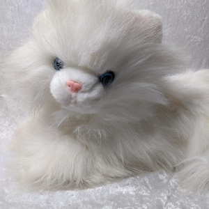 May include: A plush white cat with fluffy fur, blue eyes, and a pink nose. The stuffed animal is lying down, with its head and front paws visible. The cat's fur is soft and textured, giving it a realistic appearance.