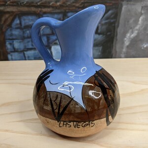 May include: A blue and brown ceramic pitcher with a handle. The pitcher has a painted design of a desert scene with the words "LAS VEGAS" on the bottom.