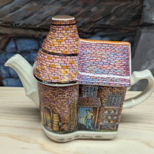 May include: A ceramic teapot shaped like a building with a brick-patterned exterior, a chimney-style lid, and a cream-colored handle and spout. The facade features a painted scene of a person standing near a doorway, windows, and shelves.