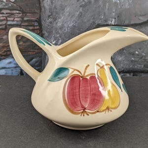 May include: A white ceramic pitcher with a curved spout and a handle. The pitcher is decorated with a hand-painted design of a red apple and yellow pears with green leaves.