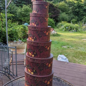 May include: A stack of seven round, burgundy-colored decorative boxes with lids. Each box is adorned with a pattern of fruits and foliage in shades of yellow, orange, and green. The boxes are arranged in descending order by size.