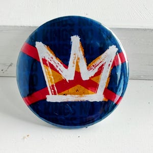 May include: A round, blue button with a white crown design. The crown is outlined in white and has red and yellow accents. Red stripes cross the button. The button is likely a pin or badge.