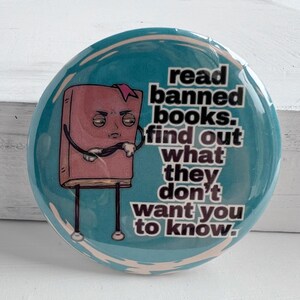 May include: A circular teal badge with the text "read banned books. find out what they don't want you to know." A cartoon book with a pink cover and a sad face is featured on the left side of the badge.