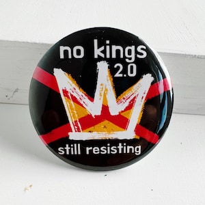 Könnte beinhalten: Ein runder, schwarzer Button mit dem weißen Text "no kings 2.0". Eine weiße Krone mit roten und gelben Akzenten ist darunter zentriert. Die Worte "still resisting" stehen am unteren Rand des Buttons.