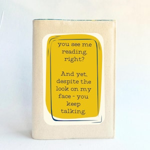 May include: A book cover with a yellow rectangle containing the text "you see me reading, right? And yet, despite the look on my face - you keep talking." The book cover is a light beige color.