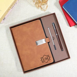 Puede incluir: Un diario de cuero marrón con un cierre plateado, presentado en una caja de regalo con un bolígrafo y un lápiz óptico a juego. El diario está grabado con las iniciales "GM" dentro de un diseño en forma de diamante. Se ven cuadernos rojos y azules adicionales.