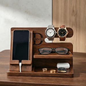 Può includere: Una docking station in legno marrone scuro dotata di supporto per smartphone con cavo di ricarica, un espositore per due orologi da polso, un ripiano per occhiali e un vassoio inferiore contenente due anelli dorati, una custodia per auricolari bianca e una chiave.