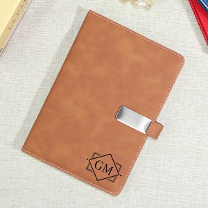 May include: A tan leather journal with a silver metal clasp. The journal has stitched edges and a geometric design with the initials "GM" engraved on the front. The journal is closed and resting on a neutral surface.