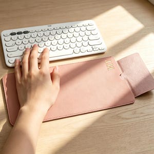 以下が含まれることがあります： 角が丸くステッチが施された長方形のピンク色のデスクマット。右上には金色のエンボス加工で「HT」のイニシャルが刻印され、右側にはフラップが付いています。丸いキーを備えた白いワイヤレスキーボードの下に配置されています。
