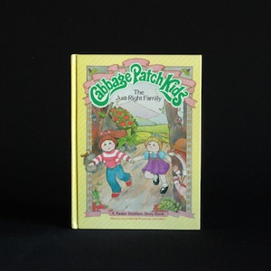 May include: A yellow book with a green and white illustration of two Cabbage Patch Kids dolls on the cover. The title of the book is "Cabbage Patch Kids The Just-Right Family" and it is a Parker Brothers Story Book.