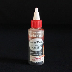 May include: A clear plastic bottle of Chefmaster Liquid Paste instant food color. The bottle has a red cap and a white tip. The label reads "Orange" and "Net Weight 2 oz."