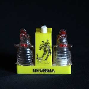 May include: A yellow salt and pepper shaker set shaped like alligators with red bow ties. The shakers are on a yellow base with a palm tree design and the word "GEORGIA" printed on the front.