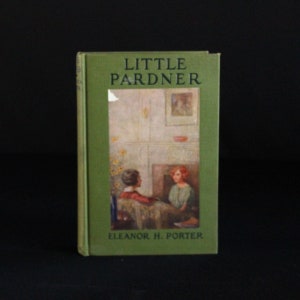 Op de afbeelding: Een groen boek met de titel "LITTLE PARNDER" en de auteur "ELEANOR H. PORTER" op de omslag. De omslag toont een illustratie van twee meisjes die bij een open haard zitten.