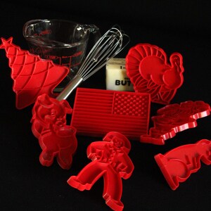 Puede incluir: Un juego de cortadores de galletas de plástico rojo en varias formas, incluyendo un pavo, un cerdo, un árbol de Navidad, una estrella, una bandera estadounidense y un hombre de jengibre. Los cortadores están sobre una superficie negra con un batidor, una taza medidora y una barra de mantequilla.