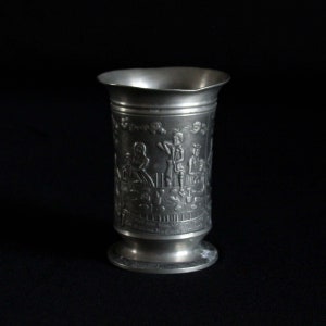 May include: A silver pewter tankard with an embossed design of people gathered around a table. The tankard has a rounded base and a flared lip.