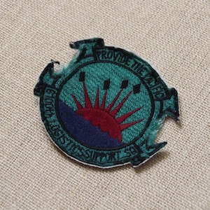 Op de afbeelding: Een geborduurde patch in groen en blauw met een rode zon die opkomt boven een blauwe en paarse bergketen. De tekst "Provide the Traffic" staat in een cirkel rond de afbeelding.