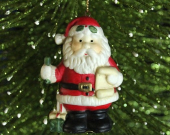 Puede incluir: Un adorno navideño de cerámica que representa a Papá Noel sosteniendo un regalo y una carta. Lleva un traje rojo con ribetes blancos, botas negras y una barba blanca.