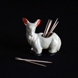 Pode incluir: Figurine de veado de cerâmica branca com orelhas vermelhas, usada como porta-palitos. Os palitos são visíveis dentro do porta-palitos.