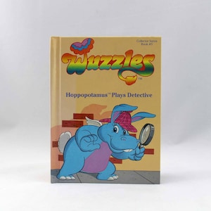 Könnte beinhalten: Ein Buch mit dem Titel "Wuggles" und dem Untertitel "Hippopotamus spielt Detektiv". Der Buchumschlag zeigt ein blaues, cartoonartiges Flusspferd, das einen rosa Hut trägt und eine Lupe hält. Das Buch ist Teil einer Sammlerserie, Buch Nummer 5.