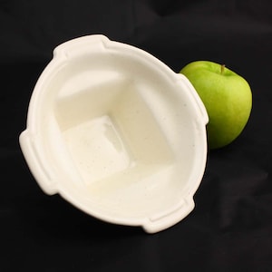 May include: A white ceramic bowl with a square-shaped interior and four handles. The bowl is empty and has a speckled texture.