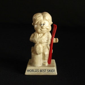 May include: A figurine of a cartoon skier with a broken leg, sitting on a log with a ski pole. The base of the figurine reads "WORLD'S BEST SKIER".