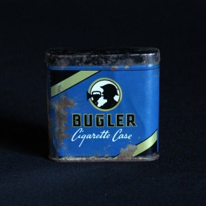 Puede incluir: Una caja de metal para cigarrillos azul y amarilla con una imagen en blanco y negro de un trompetista tocando la trompeta. La caja dice "Bugler Cigarette Case" en la parte delantera.