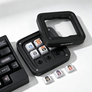 Könnte beinhalten: Ein schwarzes mechanisches Makro-Pad mit abnehmbarem Rahmen und der Aufschrift YOUR CUSTOM TEXT. Das Gerät enthält ein Raster aus weißen Tastenkappen mit bunten Charakterillustrationen, die teilweise installiert und teilweise separat platziert sind.