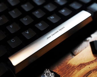 Pode incluir: Close-up de um teclado personalizado com uma barra de espaço prateada. A barra de espaço tem o texto "Your Custom Space" gravado. Uma tecla preta com um gráfico branco também é visível. O teclado está sobre uma superfície de madeira.