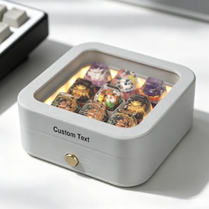 May include: A square, light grey display box with a glass top and a gold-toned circular button on the front. The text "Custom Text" is printed on the side. Inside, nine translucent, multi-coloured resin keycaps are illuminated by a warm internal light.
