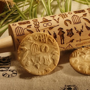 May include: A wooden rolling pin with a design of various Harry Potter themed symbols and characters. The rolling pin is on a beige linen tablecloth with two cookies that have been rolled with the pin. The cookies are in the shape of a circle and have a design of a birdcage and other symbols.
