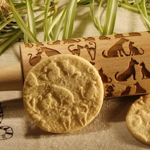 Può includere: Un mattarello di legno con un designo di gatto inciso sulla superficie. Il mattarello è su una tovaglia di lino con un motivo di gatti. Due biscotti con disegni di gatti sono accanto al mattarello.