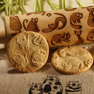 Op de afbeelding: Een houten deegroller met kattenontwerpen, samen met drie katvormige koekjes. De deegroller is lichtbruin en de koekjes zijn goudbruin. De achtergrond omvat een pot honing, een honingdipper en een patroonstof.