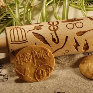 May include: A wooden rolling pin featuring a Harry Potter design. The rolling pin is engraved with various Harry Potter symbols, including the Hogwarts crest, a lightning bolt, a broom, and a pair of glasses. There are two cookies next to the rolling pin, one with the Harry Potter logo and the other with the number 9 3/4.
