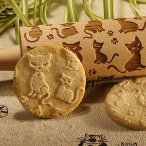 Può includere: Un mattarello di legno con un design a forma di gatto è mostrato su una tovaglia di lino beige. Il mattarello viene utilizzato per creare biscotti a forma di gatto. Due biscotti sono mostrati sulla tovaglia, uno con un design a forma di gatto e l'altro semplice. Un barattolo di miele e un mestolo per il miele sono anche mostrati nell'immagine.