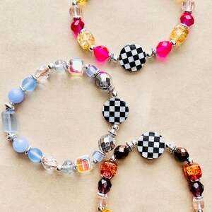 May include: Three beaded bracelets with a variety of colorful beads, including pink, yellow, blue, and brown. Each bracelet features a black and white checkered disc as a focal point, with silver-toned metal accents.