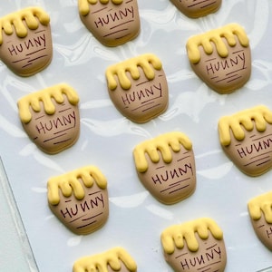 May include: A collection of small, tan-colored honey pot-shaped charms, each featuring a yellow, dripping honey design at the top and the word "HUNNY" printed in dark brown lettering, arranged in a grid pattern on a white backing sheet.