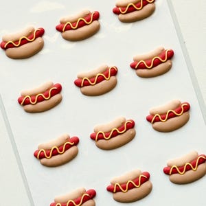 May include: A sheet of twelve small, three-dimensional hot dog embellishments. Each piece features a tan bun, a red sausage, and a yellow wavy line of mustard. The hot dogs are arranged in a grid pattern on a smooth, white, semi-transparent backing.