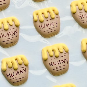 May include: A collection of tan-colored cookies shaped like honey pots, each topped with yellow icing dripping down the sides. The word HUNNY is written in dark brown icing on the front of each pot, accompanied by horizontal lines below the text.