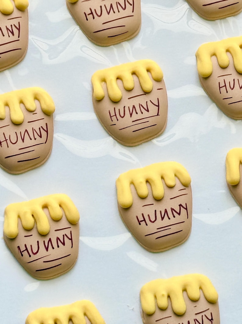 May include: A collection of tan-colored cookies shaped like honey pots, each topped with yellow icing dripping down the sides. The word HUNNY is written in dark brown icing on the front of each pot, accompanied by horizontal lines below the text.