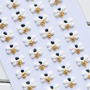 May include: A sheet of small, three-dimensional bee-shaped embellishments arranged in a grid. Each bee features white textured wings, a dark blue head, and a gold-colored body with horizontal black stripes, creating a repeating pattern.