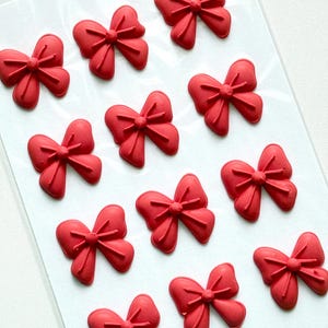 May include: A set of twelve individual red bow-shaped embellishments arranged in a grid pattern on a clear plastic sheet. Each bow features a central knot with radiating lines and two symmetrical loops on either side, creating a uniform design.