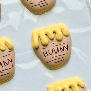 May include: Tan-colored, pot-shaped cookies decorated with yellow icing drips at the top and the word "HUNNY" written in brown script. Each cookie features horizontal brown lines below the text, mimicking the appearance of a honey pot.