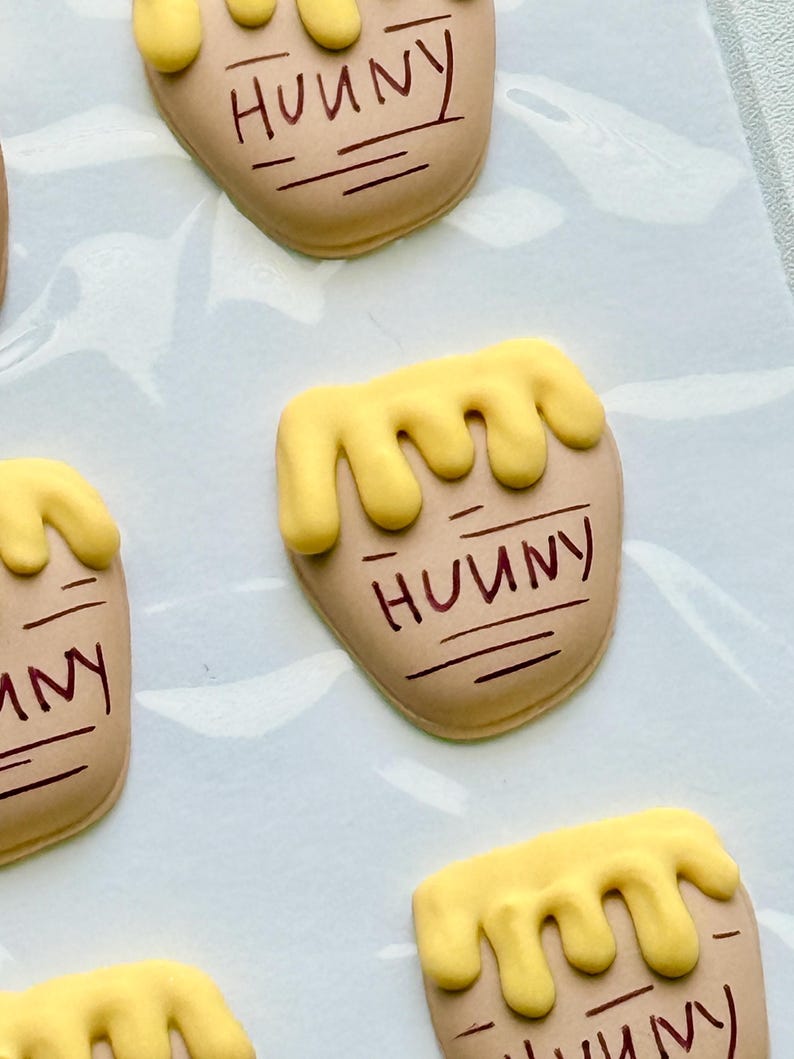 May include: Tan-colored, pot-shaped cookies decorated with yellow icing drips at the top and the word "HUNNY" written in brown script. Each cookie features horizontal brown lines below the text, mimicking the appearance of a honey pot.