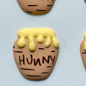 May include: Close-up of several brown, oval-shaped objects resembling honey pots. Each pot has a yellow, dripping honey design at the top and the word "Hunny" written in brown.